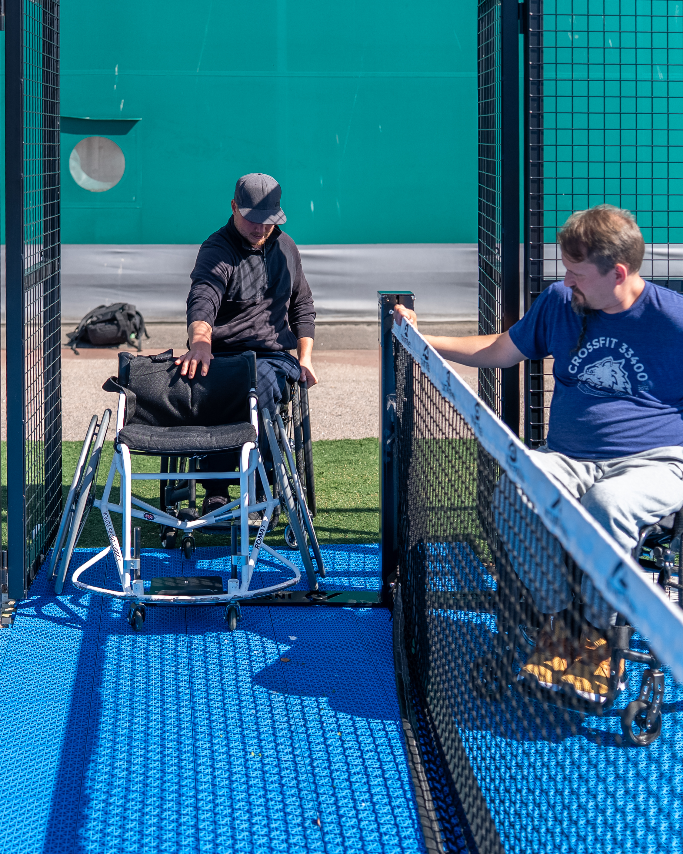 Kuvassa henkilö pyörätuolissa siirtää padelverkkoa, että toinen henkilö pääsee padelkentälle pyörätuolilla työntäen pelipyörätuolia edellään.